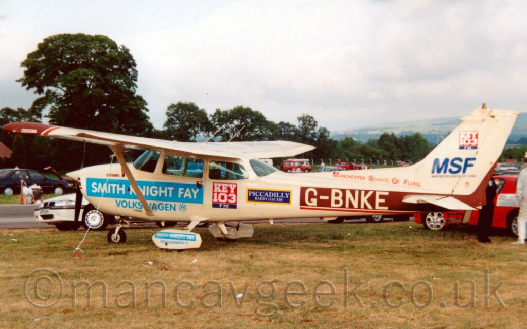 Side view of a high-winged, propellor-engined light aircraft parked facing to the left on grass. The plane is in a mostly a cream colour, with large blue "Smith Knight Fay Volkswagon" stickers on the forward fuselage under the cockpit and also on the sponsons over the main landing gear, a red square with white text "Key 103" over a blue bar with white text "FM" under the rear of the cabin, a blue bar outlined in yellow with the white text" Piccadilly Radio 1152AM", and a brown bar stretching along the rear fuselage with the cream registration "G-BNKE" on top. There is red text "Manchester School Of Flying" on a fillet on top of the fuselage, at the base of the tail, with a blue "MSF" logo and text "Manchester School Of Flying" on the base of the tail. Another red and blue square with text "Key 103 FM" is at the top of the tail. Green grass fills the foreground, with cars parked on grass in the background, and trees in the distance under grey sky.