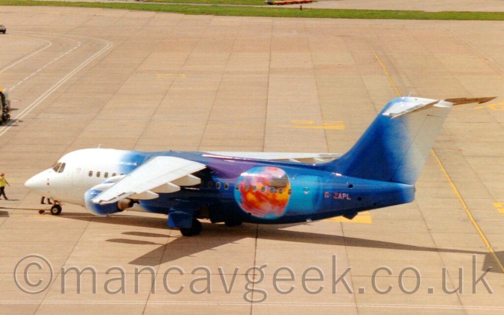 Elevated side view of a high-winged, 4 engined jet airliner facing to the left but being pushed back to the right by an out-of-frame tug attached to the nose wheel by a long yellow pole. The planes forward fuselage, wings, and the upper-rear of the tail are white, the rest filled with a depiction of space in blue, black, and purple, with an orange, green, red, and blue planet on the rear fuselage, next to the multi-coloured registration "G-ZAPL". Square panels of grey concrete apron fill the foreground, as well as most of the background, with a strip of grass at the top of the image.