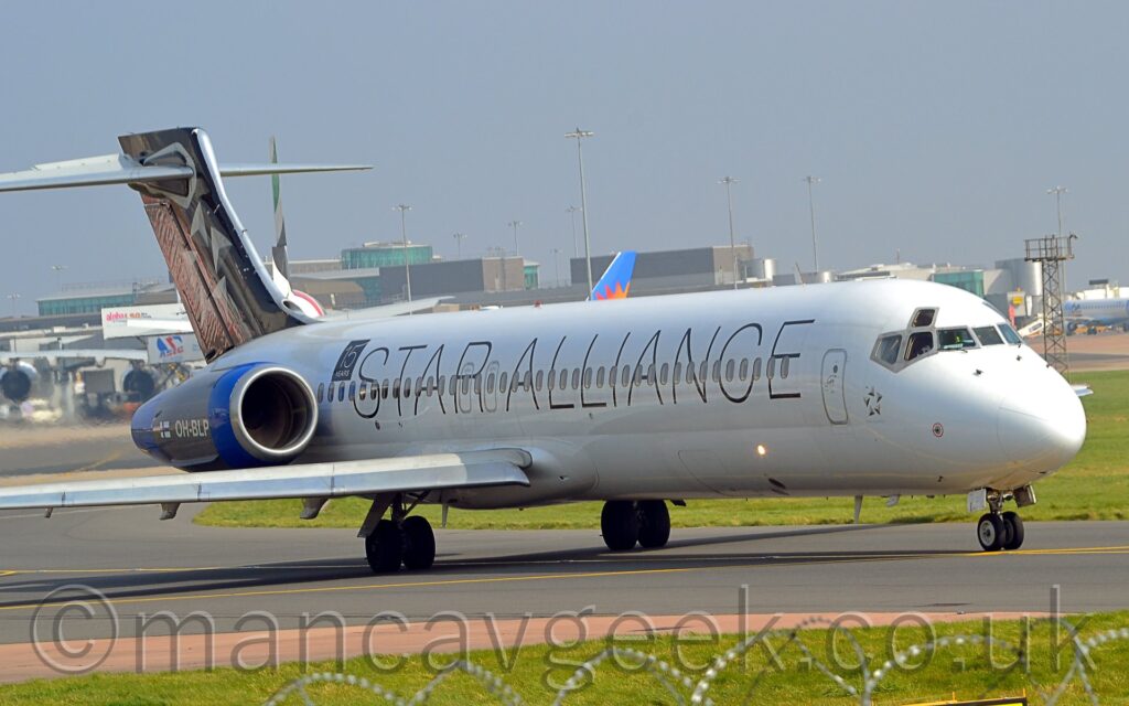 Side view of a twin engined jet airliner with the engines mounted on the sides of the rear fuselage, taxiing from left to right along a grey taxiway. The plane is mostly white, with billboard-style "Star Alliance" titles on the side of the fuselage, running the length of the body. The engine pods on the rear fuselage are blue, with white registration "OH-BLP" next to a small Finnish flag on the side, with a black square with white text "15 years" on the upper rear fuselage just in front. The black tail has a circle of 5 white and grey arrowheads pointing in different directions. Green grass lines the sides of the taxiway in the foreground, with a grey terminal building stretching across the frame from the left, with several planes parked in front, under pale blue-grey sky.