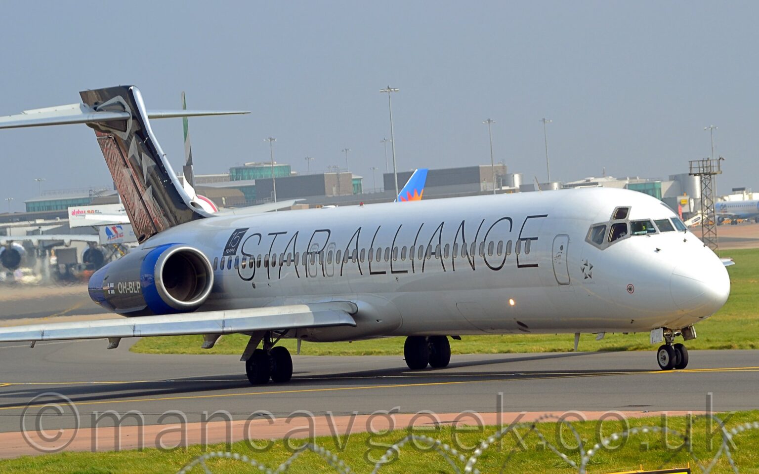 Side view of a twin engined jet airliner with the engines mounted on the sides of the rear fuselage, taxiing from left to right along a grey taxiway. The plane is mostly white, with billboard-style "Star Alliance" titles on the side of the fuselage, running the length of the body. The engine pods on the rear fuselage are blue, with white registration "OH-BLP" next to a small Finnish flag on the side, with a black square with white text "15 years" on the upper rear fuselage just in front. The black tail has a circle of 5 white and grey arrowheads pointing in different directions. Green grass lines the sides of the taxiway in the foreground, with a grey terminal building stretching across the frame from the left, with several planes parked in front, under pale blue-grey sky.