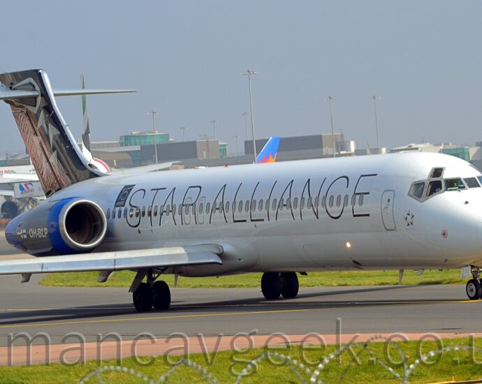 Side view of a twin engined jet airliner with the engines mounted on the sides of the rear fuselage, taxiing from left to right along a grey taxiway. The plane is mostly white, with billboard-style "Star Alliance" titles on the side of the fuselage, running the length of the body. The engine pods on the rear fuselage are blue, with white registration "OH-BLP" next to a small Finnish flag on the side, with a black square with white text "15 years" on the upper rear fuselage just in front. The black tail has a circle of 5 white and grey arrowheads pointing in different directions. Green grass lines the sides of the taxiway in the foreground, with a grey terminal building stretching across the frame from the left, with several planes parked in front, under pale blue-grey sky.