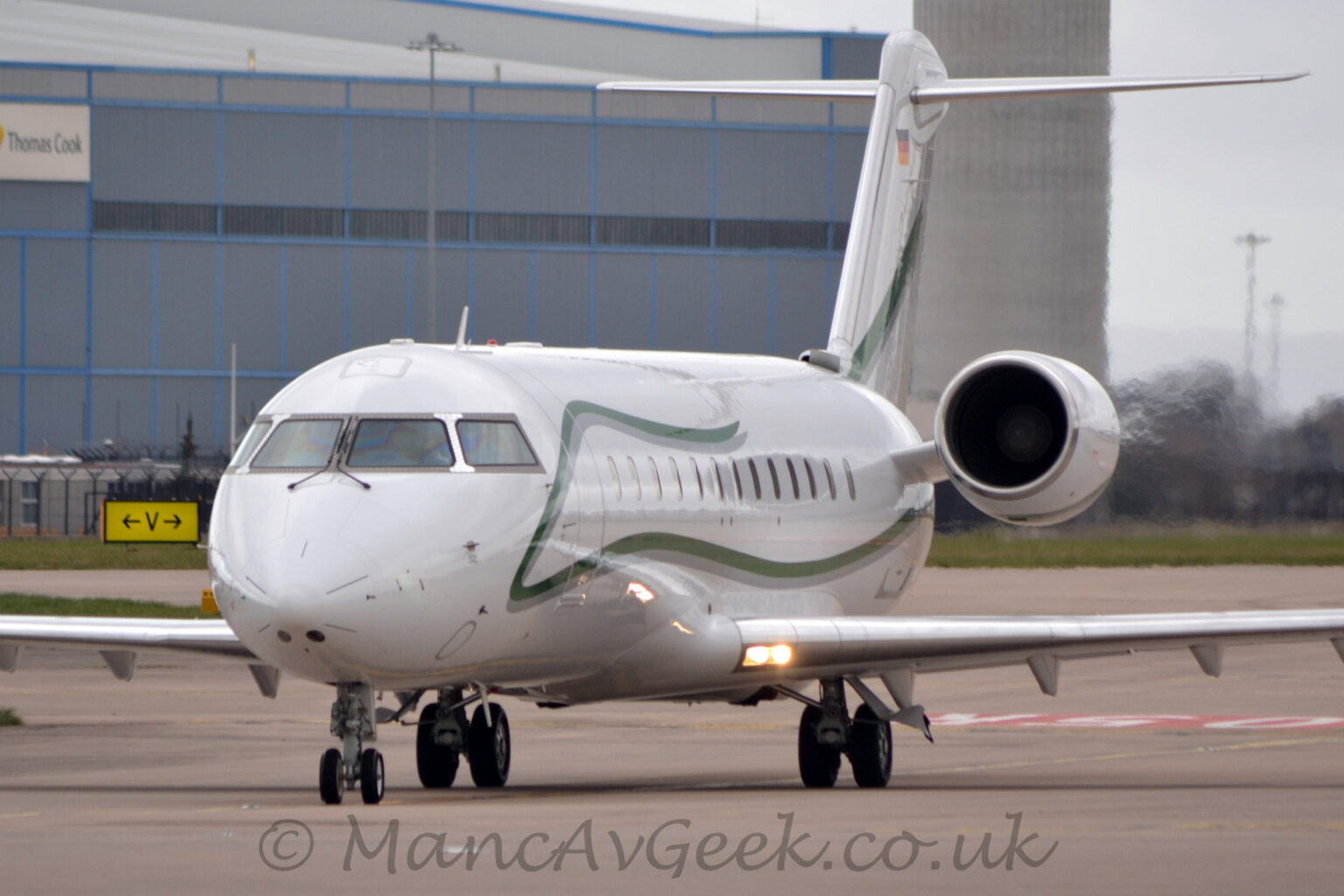 Front view of a twin engined BizJet taxiing from right to left towards the camera on a wide grey taxiway. The plane is mostly white, with a green squiggle with a grey drop shadow running from under the engines mounted on the sides of the rear fuselage, forwards along the body under the passenger cabin window, then up between the cockpit windows and the main door, then rearwards again, over the top of the cabin windows, stopping about half-way along the body S similar strip runs from the bottom left to top right of the tail. There are a pair of bright lights shining in the wing root, creating a slight flare in the image, and causing bright reflections on the fuselage. A large grey and blue hangar dominates the background on the left, with cloudy grey sky filling the rest of the frame.