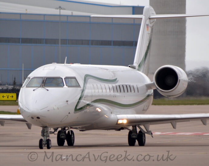 Front view of a twin engined BizJet taxiing from right to left towards the camera on a wide grey taxiway. The plane is mostly white, with a green squiggle with a grey drop shadow running from under the engines mounted on the sides of the rear fuselage, forwards along the body under the passenger cabin window, then up between the cockpit windows and the main door, then rearwards again, over the top of the cabin windows, stopping about half-way along the body S similar strip runs from the bottom left to top right of the tail. There are a pair of bright lights shining in the wing root, creating a slight flare in the image, and causing bright reflections on the fuselage. A large grey and blue hangar dominates the background on the left, with cloudy grey sky filling the rest of the frame.