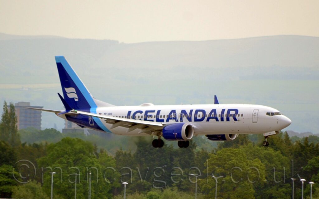 Side view of a white, twin engined jet airliner flying from left to right at a very low altitude, with undercarriage lowered and flaps extended from the rear of the wings. There are large, billboard-style, dark blue "Icelandair" titles on the forward fuselage, with dark blue engine pods in front of and below the wings, and a dark blue tail and rear fuselage. There 2 thick, white, waving stripes on the tail, with a pale blue diagonal stripe running down the front of the tail and across the rear fuselage. Trees in the distance fill the bottom of the frame, with cloudy grey skies filling the top of the frame.