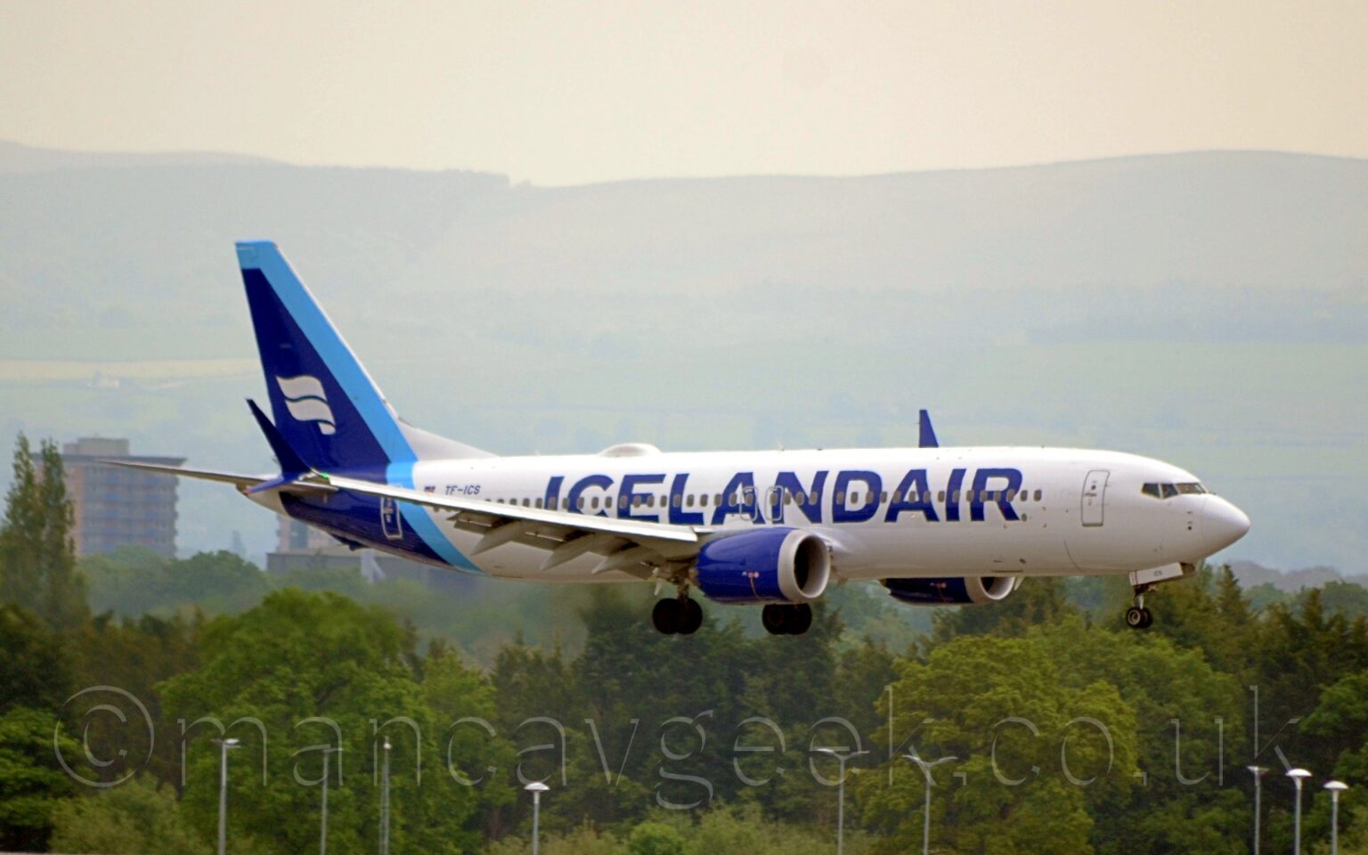 Side view of a white, twin engined jet airliner flying from left to right at a very low altitude, with undercarriage lowered and flaps extended from the rear of the wings. There are large, billboard-style, dark blue "Icelandair" titles on the forward fuselage, with dark blue engine pods in front of and below the wings, and a dark blue tail and rear fuselage. There 2 thick, white, waving stripes on the tail, with a pale blue diagonal stripe running down the front of the tail and across the rear fuselage. Trees in the distance fill the bottom of the frame, with cloudy grey skies filling the top of the frame.