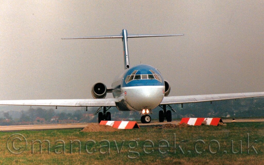 Almost head-on view from slightly below of a blue and grey, twin engined jet airliner with the engines mounted on the sides of the rear fuselage, turning to it's right as it lines up on a runway. The upper body is a dark blue, with a grey belly, a white stripe running along the body separating the 2 sections. A small British flag is visible on each side of the lower forward fuselage, under the rear cockpit windows. Green grass fills the foreground, with piles of dirt on the ground, with red and white striped triangular frames making their location. Under the planes wings, trees and houses can be seen in the distance, with dismal grey sky filling the rest of the frame.