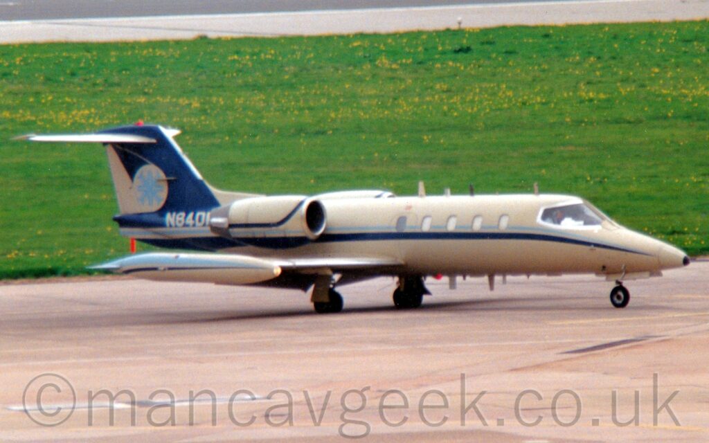 Side view of a twin engined BizJet with the engines mounted on the sides of the rear fuselage, parked on grey concrete apron marked with faded yellow hash markings. The plane is largely a sandy brown, with a dark and light blue stripe running along the body under the passenger cabin windows and along the bottom of the brown engine pods, and the top of the wing-tip fuel tanks. The tail is dark blue, with the white registration "N84DM" at the bottom, and a sandy brown circle containing something that at first glance looks like a pale blue snowflake in the middle. Green grass littered with small yellow flowers fills the background, with a black runway right at the top.