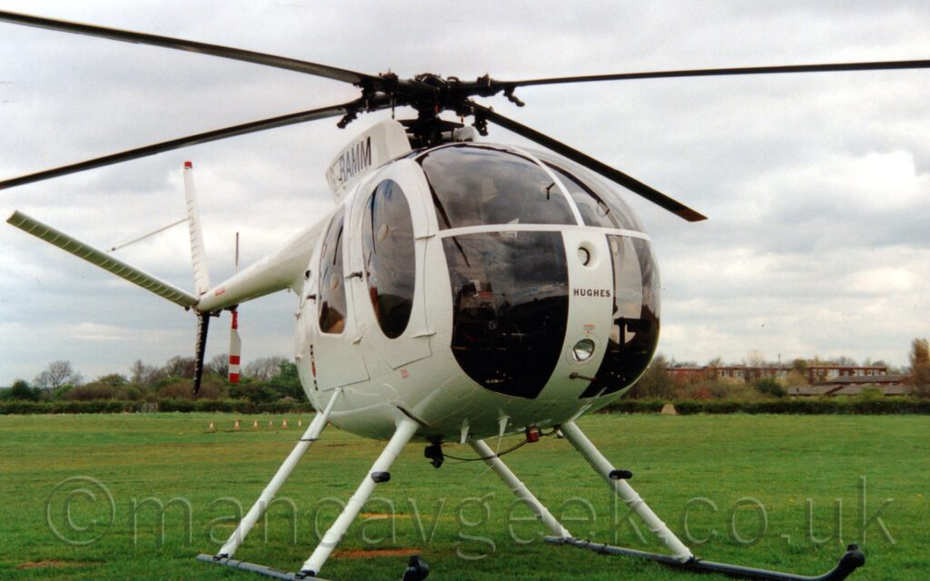 Front view of a white helicopter with a heavily glazed nose parked on grass. The black word "Hughes" is on a white metal panel at the front, and the black registration "G-RAMM" on the sides of the engine pod, mounted on top of the fuselage. Green grass fills the bottom half of the frame in the background, with hedges beyond that, and brown houses behind those, under streaky white clouds with hints of blue poking through.