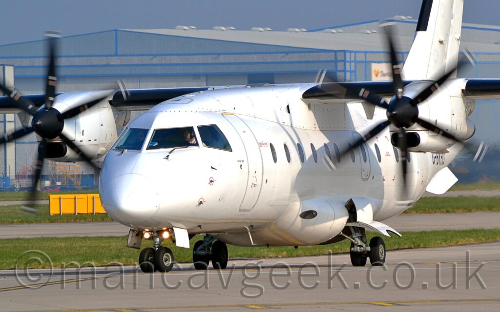 Side view of a high-winged, twin propellor-engined airliner taxiing from right to left. The plane is almost entirely white, with only small airline and type logos either side of the forward door, doors outlined in silver, and emergency exits marked in red to break the monotony. The six-bladed propellors are blurred just enough to convey movement without appearing to be discs. A grey concrete runway is in the foreground, with green grass and grey concrete taxiways in the background, with a large grey hangar in the distance, under a grey-blue sky.