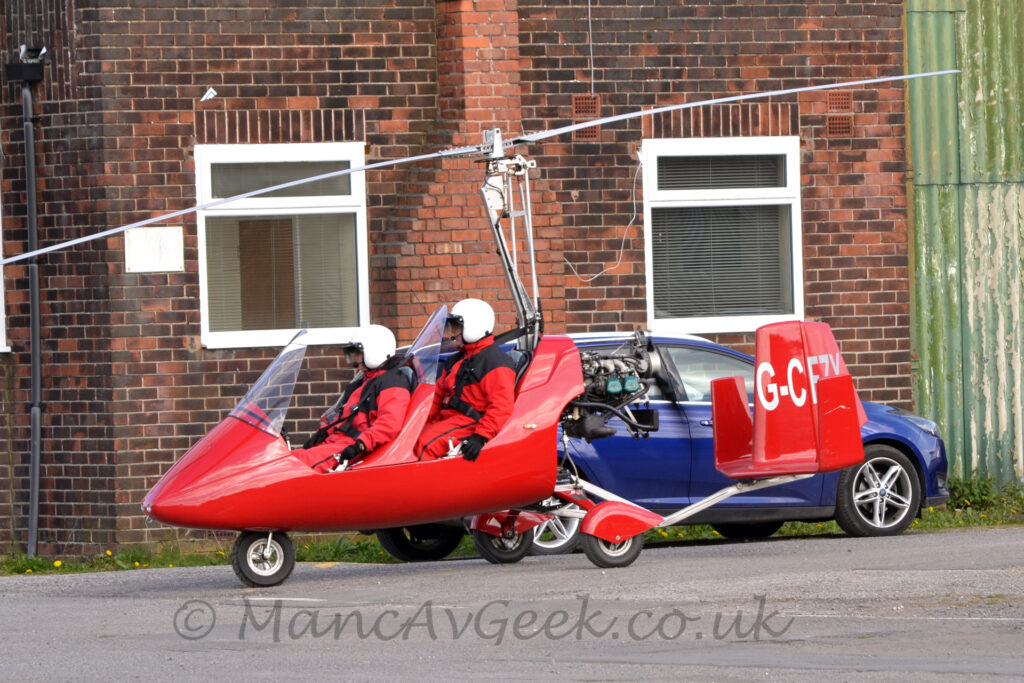 Side view of a red, 2 seat autogyro taxiing from right to left. Both seats in the tandem twin cockpits are filled with people wearing red flight suits and white helmets, secured into place by wide black straps. The red triple tail extends rearwards from under the rear-facing engine, with the white registration "G-CFZX" on the centre tail. A brown brick building fills the background, with a blue car parked in front, behind this autogyro.