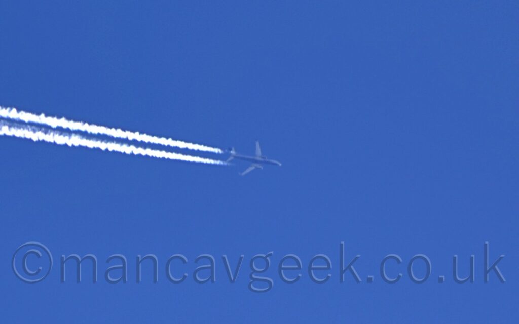 A white, twin engined jet airliner with a dark blue belly and engine pods, flying from left to right at a very high altitude, with a long plume of condensation coming from the rear of each engine. Vivid blue sky fills the rest of the frame.