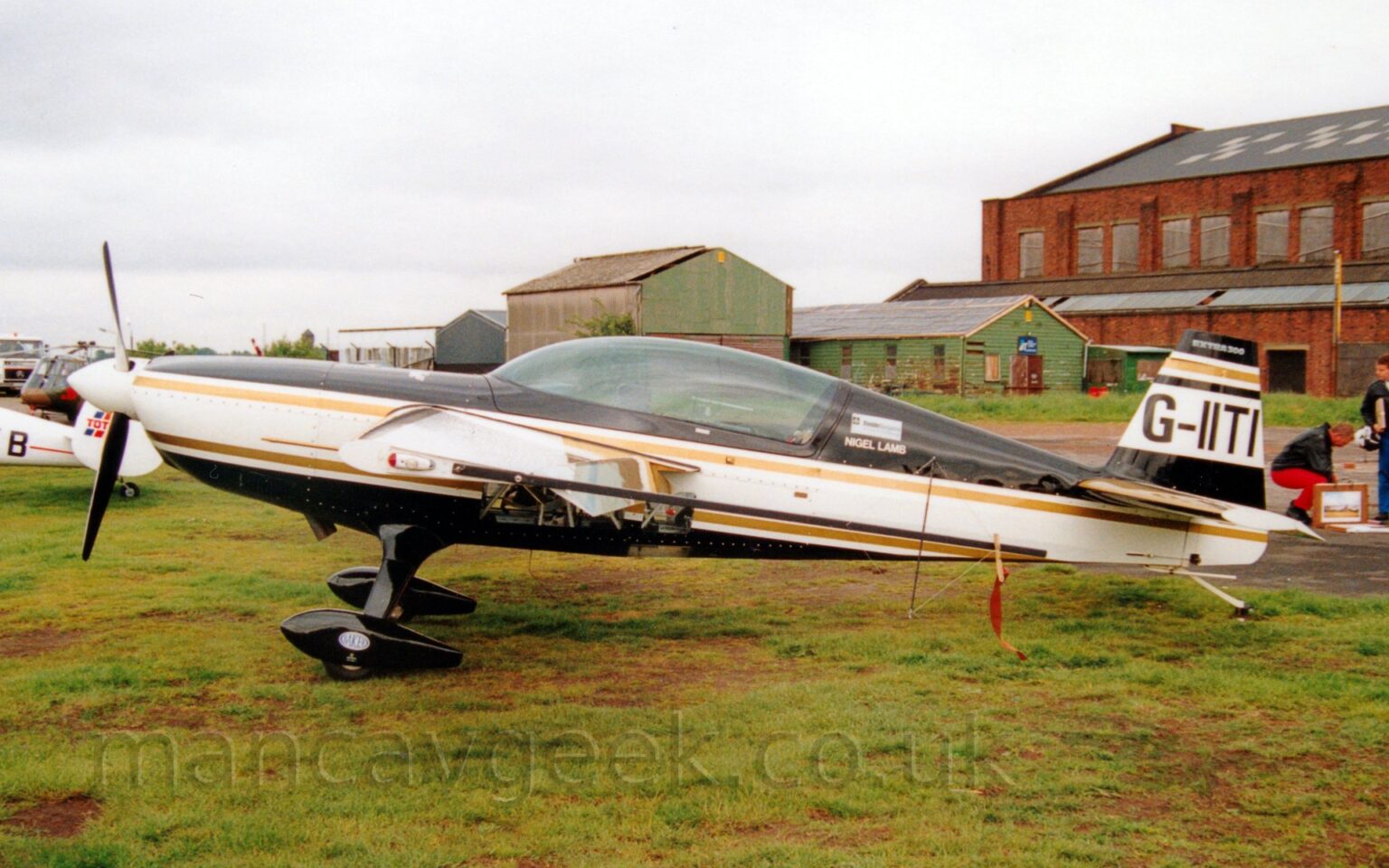 Side view of a white and black, single engined aerobatic light aircraft parked on grass facing to the left. The plane has black top and bottom surfaces, with a thick white stripe running along the body, and gold piping in between. The registration "G-IITI" is on a thick white stripe on the black tail. There is a large, glazed canopy over the cockpit, with small white "Nigel Lamb" titles on the fuselage immediately behind the canopy. There are also glazed panels in the lower fuselage, under the wings. Black spats cover the wheels on the fixed landing gear. Green grass fills the foreground. A large, red brick hangar is on the right of the frame in the background, with smaller green buildings next to it, towards the middle of the frame. The tail of a white light aircraft and a small, camouflaged helicopter are in the background on the left. Cloudy grey sky fills the rest of the frame.