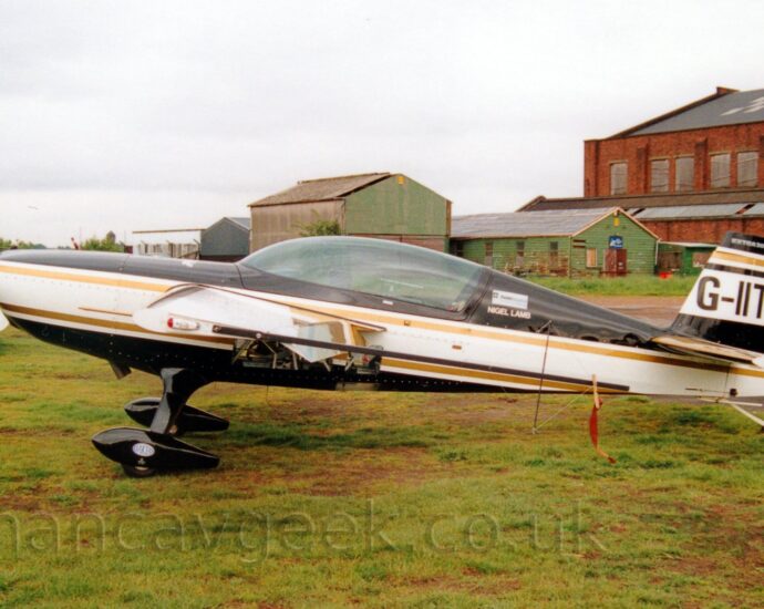 Side view of a white and black, single engined aerobatic light aircraft parked on grass facing to the left. The plane has black top and bottom surfaces, with a thick white stripe running along the body, and gold piping in between. The registration "G-IITI" is on a thick white stripe on the black tail. There is a large, glazed canopy over the cockpit, with small white "Nigel Lamb" titles on the fuselage immediately behind the canopy. There are also glazed panels in the lower fuselage, under the wings. Black spats cover the wheels on the fixed landing gear. Green grass fills the foreground. A large, red brick hangar is on the right of the frame in the background, with smaller green buildings next to it, towards the middle of the frame. The tail of a white light aircraft and a small, camouflaged helicopter are in the background on the left. Cloudy grey sky fills the rest of the frame.