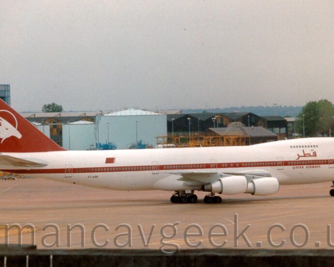 Side view of a very large, 4 engined jet airliner taxiing from left to right. The plane is mostly white, with a maroon stripe running along the body, covering the lower deck windows, and maroon "Qatar Airways" titles on the forward fuselage, in an Arabic script between the upper and lower deck windows, and in English below the lower deck windows. The tail is also maroon, with the white head of a horned, goat-like animal inside the outline of a white circle. Grey concrete apron fills the foreground, with a large grey hangar in the background on the left, several pale blue fuel tanks towards the middle, and a selection of other airport buildings on the right. Trees in the distance slowly vanish into a bright but hazy sky.