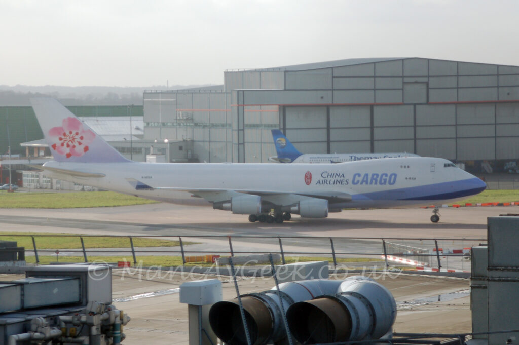 Side view of a very large, 4 engined jet airliner taxiing from left to right. The plane is mostly white, with diagonal light and blue bands around the plane's nose, with large black "China Airlines" titles on the upper forward fuselage, next to larger blue "Cargo" text. The black registration "B-18721" is under the upper deck windows, as well as on the upper rear fuselage. The tail is white at the top and a pale purple at the bottom, with a pink and red flower filling most of the rest. The roof of a terminal building fills the foreground, with large air conditioning ducts in the middle, with large grey metal structures on either side of the screen. A large grey hangar fills most of the background, with a white and blue twin engined jet airliner parked in front. Hazy grey skies fill the rest of the frame.