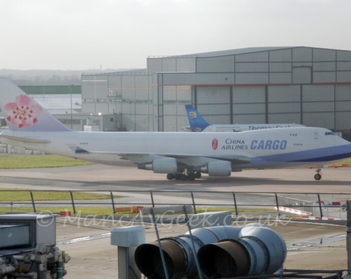 Side view of a very large, 4 engined jet airliner taxiing from left to right. The plane is mostly white, with diagonal light and blue bands around the plane's nose, with large black "China Airlines" titles on the upper forward fuselage, next to larger blue "Cargo" text. The black registration "B-18721" is under the upper deck windows, as well as on the upper rear fuselage. The tail is white at the top and a pale purple at the bottom, with a pink and red flower filling most of the rest. The roof of a terminal building fills the foreground, with large air conditioning ducts in the middle, with large grey metal structures on either side of the screen. A large grey hangar fills most of the background, with a white and blue twin engined jet airliner parked in front. Hazy grey skies fill the rest of the frame.