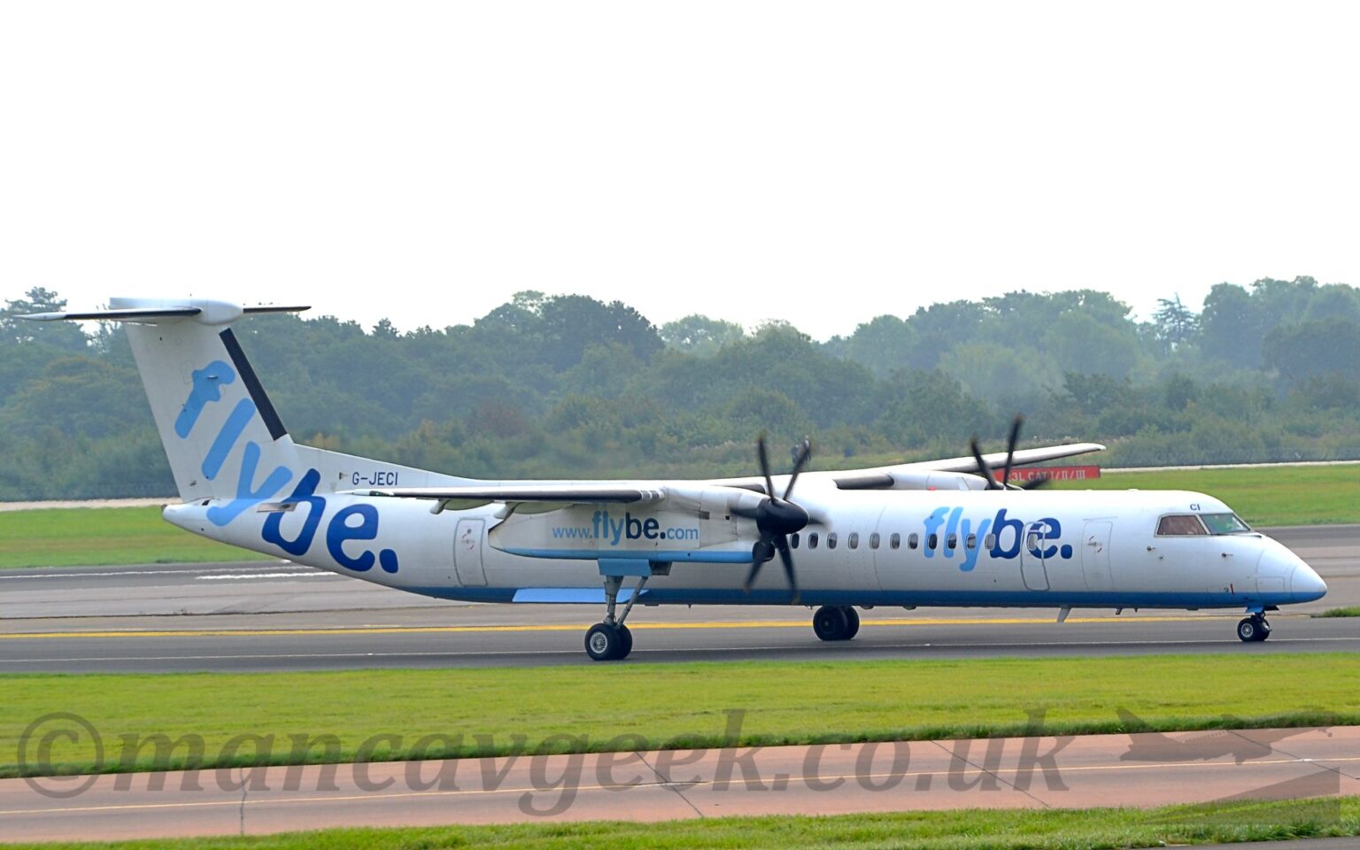 Side view of a white, high-winged, twin propellor=engined airliner taxiing from left to right along a black taxiway. The plane is mostly white, with a pale blue belly, and light and dark blue "FlyBe" titles on the side of the forward fuselage. A larger version of this logo is on the white tail and rear fuselage, and there is the blue web address "www[dot]flybe[dot]com" on the sides of the engine pods, mounted under each wing. Strips of green grass line another taxiway in the foreground, with more grass in the background leading up to trees in the distance, under bright but hazy sky.