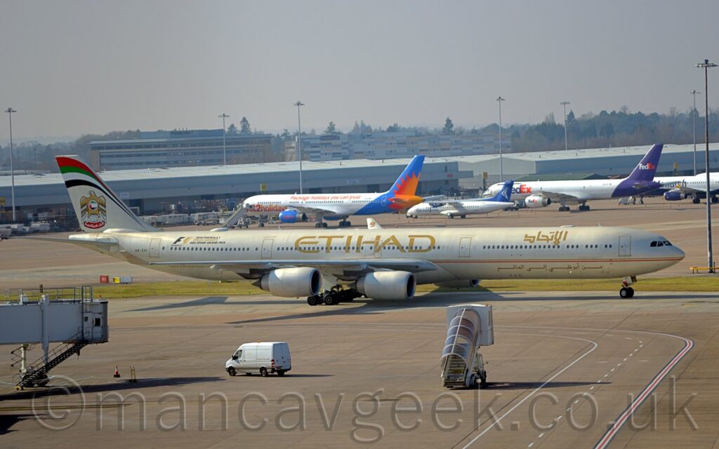Side view of a long, thin, 4 engined jet airliner taxiing from left to right. The plane is in a pale cream colour, with a gold and red pinstripe running along the body, under the passenger cabin windows. There are large, gold, "Etihad" titles on the upper centre fuselage, with a smaller version in an Arabic script on the upper forward fuselage. There is more Arabic text, in gold, on the lower forward fuselage. There is a large sticker with black and gold text on the upper rear fuselage, with a large black and gold "F1", then smaller text "2016 Formula 1 Etihad Airways Abu Dhabi Grand Prix". Grey concrete apron fills the foreground, with a mobile set of airstairs with a brown perspex awning over the top seeming to drive towards this plane, and a white van driving from right to left by an airbridge on the left of the frame that seems to hang out of nowhere. Low, grey cargo sheds stretch across the background, with several planes parked nose-in towards them. Taller office buildings and hotels nestled in trees are in the distance, under hazy grey sky.