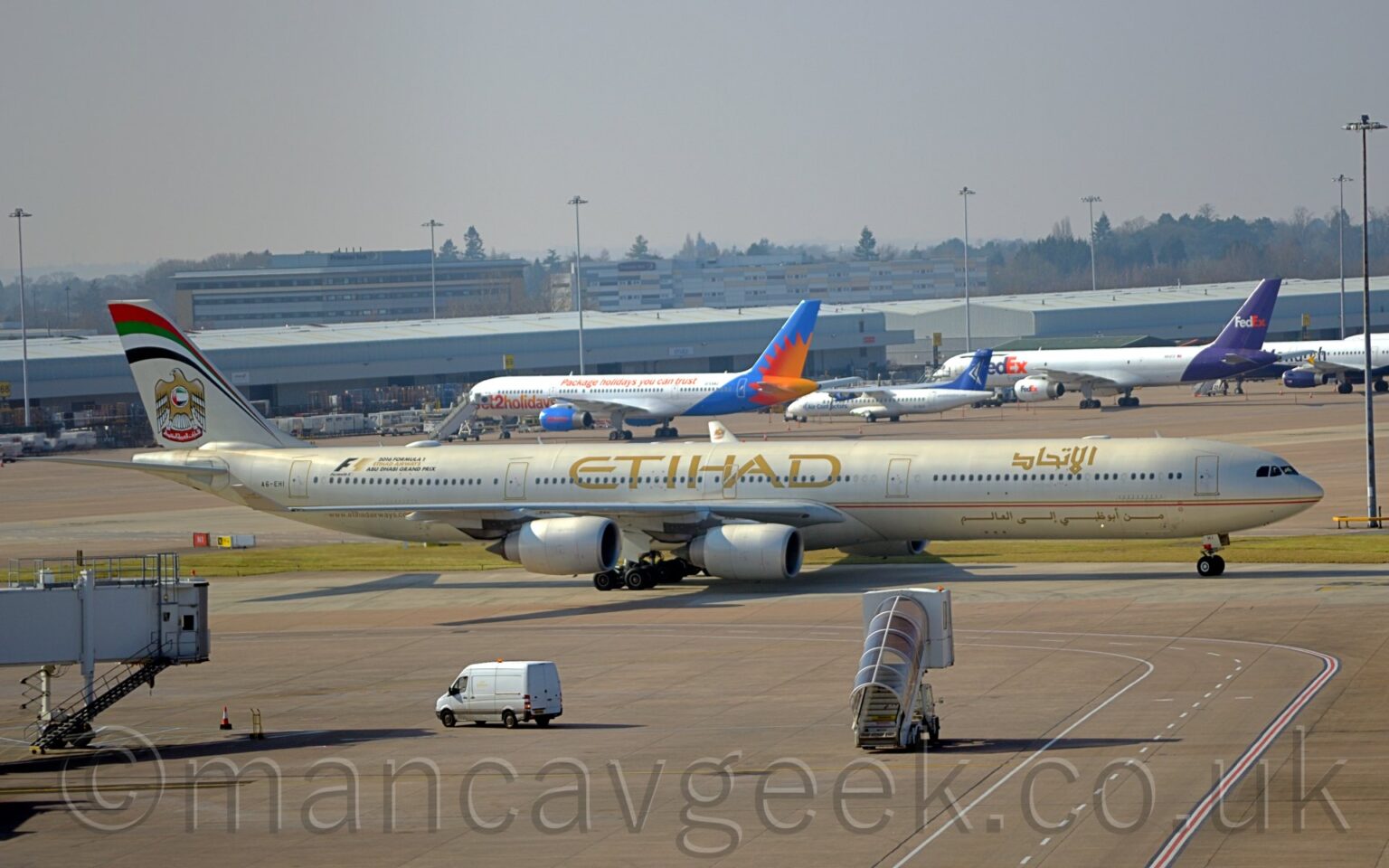 Side view of a long, thin, 4 engined jet airliner taxiing from left to right. The plane is in a pale cream colour, with a gold and red pinstripe running along the body, under the passenger cabin windows. There are large, gold, "Etihad" titles on the upper centre fuselage, with a smaller version in an Arabic script on the upper forward fuselage. There is more Arabic text, in gold, on the lower forward fuselage. There is a large sticker with black and gold text on the upper rear fuselage, with a large black and gold "F1", then smaller text "2016 Formula 1 Etihad Airways Abu Dhabi Grand Prix". Grey concrete apron fills the foreground, with a mobile set of airstairs with a brown perspex awning over the top seeming to drive towards this plane, and a white van driving from right to left by an airbridge on the left of the frame that seems to hang out of nowhere. Low, grey cargo sheds stretch across the background, with several planes parked nose-in towards them. Taller office buildings and hotels nestled in trees are in the distance, under hazy grey sky.