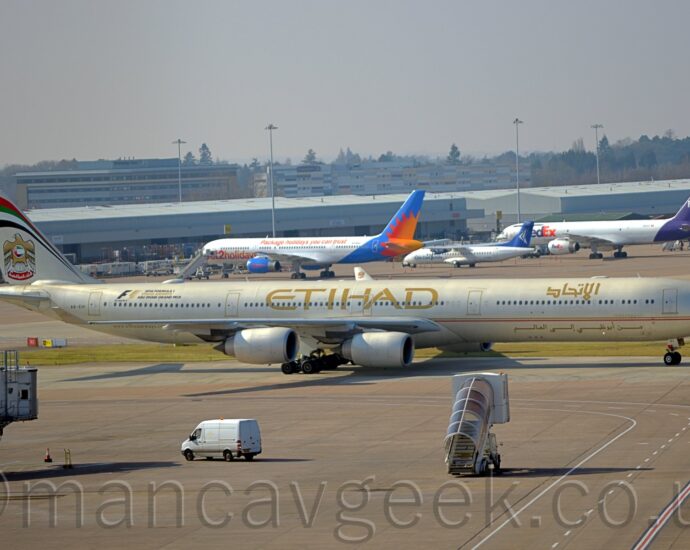 Side view of a long, thin, 4 engined jet airliner taxiing from left to right. The plane is in a pale cream colour, with a gold and red pinstripe running along the body, under the passenger cabin windows. There are large, gold, "Etihad" titles on the upper centre fuselage, with a smaller version in an Arabic script on the upper forward fuselage. There is more Arabic text, in gold, on the lower forward fuselage. There is a large sticker with black and gold text on the upper rear fuselage, with a large black and gold "F1", then smaller text "2016 Formula 1 Etihad Airways Abu Dhabi Grand Prix". Grey concrete apron fills the foreground, with a mobile set of airstairs with a brown perspex awning over the top seeming to drive towards this plane, and a white van driving from right to left by an airbridge on the left of the frame that seems to hang out of nowhere. Low, grey cargo sheds stretch across the background, with several planes parked nose-in towards them. Taller office buildings and hotels nestled in trees are in the distance, under hazy grey sky.