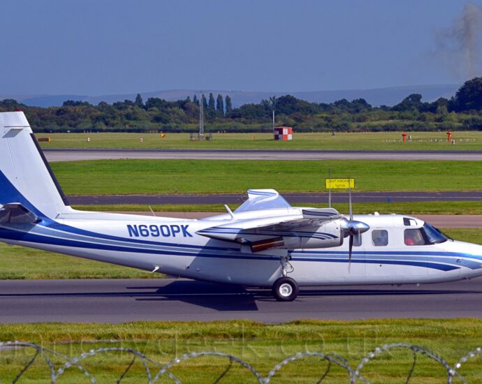 Side view of a high-winged, twin engined BizProp taxiing from left to right. The plane is largely white, with a 2-tone blue stripe running along the body from under the nose, and sweeping up into the lower parts of the tail. The registration N690PK is on the rear fuselage. Green grass fills the bottom of the frame, lining each side of the taxiway, with more grass in the background, leading up to trees in the distance under blue sky.