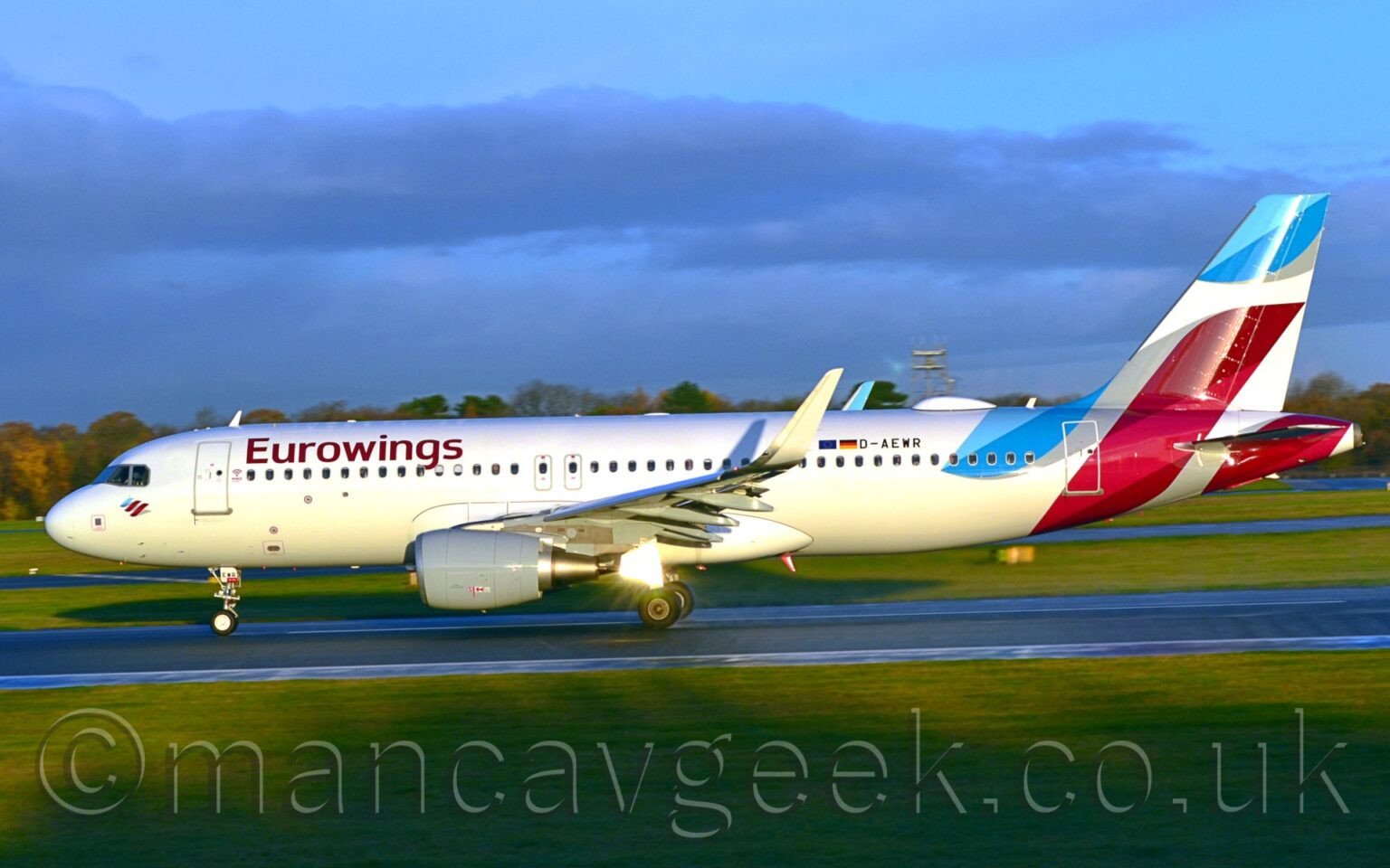 Side view of a twin engined jet airliner moving at speed from right to left along a black runway. The plane is largely white, with purple "EuroWings" titles on the upper forward fuselage, and smaller, black registration "D-AEWR" just aft of small EU and German flags on the upper rear. Pale blue and purple swirls adorn the tail and tail cone, some connected by grey swirls. Bright sunlight reflects strongly off one of the open landing gear doors under the wing, suggesting that the sun is more or less behind the camera. Green grass fills the foreground, lining the side of the runway, with more grass visible under the plane in the background, with trees in the distance under pale blue sky with purple fluffs of cloud.