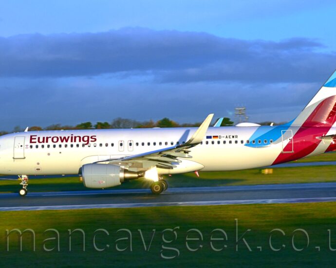 Side view of a twin engined jet airliner moving at speed from right to left along a black runway. The plane is largely white, with purple "EuroWings" titles on the upper forward fuselage, and smaller, black registration "D-AEWR" just aft of small EU and German flags on the upper rear. Pale blue and purple swirls adorn the tail and tail cone, some connected by grey swirls. Bright sunlight reflects strongly off one of the open landing gear doors under the wing, suggesting that the sun is more or less behind the camera. Green grass fills the foreground, lining the side of the runway, with more grass visible under the plane in the background, with trees in the distance under pale blue sky with purple fluffs of cloud.