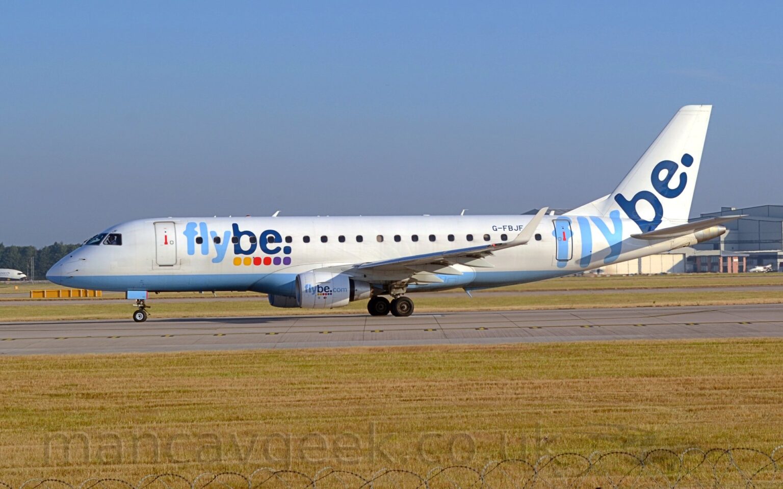 Side view of a white, twin engined jet airliner facing to the left on a grey runway. The plane has a pale blue belly, and pale and dark blue "FlyBe" titles above a series of yellow, red, and purple dots on the forward fuselage. These titles are repeated on the sides of the engine cowlings with additional "[dot] com" titles, as well as on the tail, minus the dots. Green grass fills the bottom of the frame, with the razor-wire on the top of the perimeter fence visible right at the bottom. More grass in the background leads up to a large grey hangar in the distance on the right, and trees on the left, under blue-grey sky.