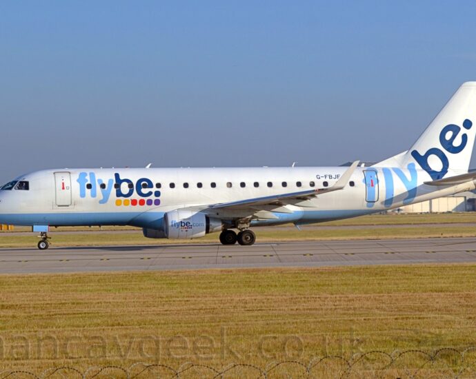 Side view of a white, twin engined jet airliner facing to the left on a grey runway. The plane has a pale blue belly, and pale and dark blue "FlyBe" titles above a series of yellow, red, and purple dots on the forward fuselage. These titles are repeated on the sides of the engine cowlings with additional "[dot] com" titles, as well as on the tail, minus the dots. Green grass fills the bottom of the frame, with the razor-wire on the top of the perimeter fence visible right at the bottom. More grass in the background leads up to a large grey hangar in the distance on the right, and trees on the left, under blue-grey sky.