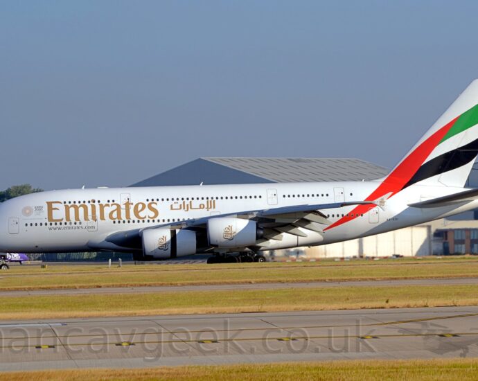 Side view of a very large, 4 engined jet airliner moving from right to left along a runway with the rear panel of one of the engines slid back, exposing the thrust reversers, suggesting it has just landed. The plane is mostly white, with large gold "Emirates" titles in English on the upper forward fuselage, between the 2 rows of passenger windows, and smaller titles in an Arabic script a bit further back, over the wings. A waving United Arab Emirates flag (red vertical bar on the left, with equal green, white, and black stripes on the right) covers the tail and part of the rear fuselage. Golden calligraphic art made up of the characters that make up the airlines Arabic name adorns the engine pods, the cowling of the inner engines split as the rear portion has moved backwards to expose the thrust reversers. Another runway stretches across the foreground, lined by green grass, while a pair of large grey hangars dominate the background, under pale blue-grey sky.