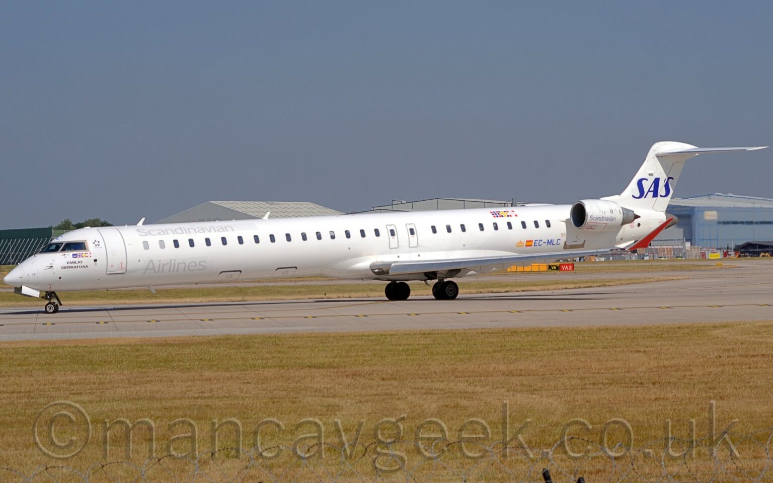 Side view of a long, thin, twin engined jet airliner taxiing from right to left as it turns on to a grey runway. The plane is mostly white, with grey "Scandinavian" titles on the upper forward fuselage, with additional "Airlines" titles on the lower forward fuselage. Smaller "Scandinavian" titles are on the sides of the white engine pods, mounted on the sides of the rear fuselage, just in front of and below the tail. The blue registration "EC-MLC" is on the lower rear fuselage, just aft of a small Spanish flag, which itself is just aft of a stylised golden crown. On the upper rear fuselage, there is a 9*3 grid of coloured dots, representing the flags of Norway, Sweden, and Denmark, the 3 countries for whom SAS is the nominal flag carrier. Slightly yellowing grass fills the foreground, along the side of the runway, while grey hangars are visible over the top of the plane in the background, under blue skies.