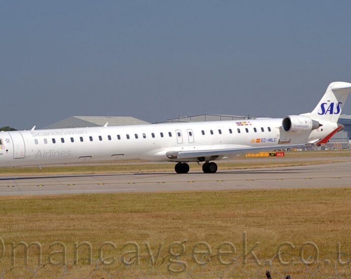 Side view of a long, thin, twin engined jet airliner taxiing from right to left as it turns on to a grey runway. The plane is mostly white, with grey "Scandinavian" titles on the upper forward fuselage, with additional "Airlines" titles on the lower forward fuselage. Smaller "Scandinavian" titles are on the sides of the white engine pods, mounted on the sides of the rear fuselage, just in front of and below the tail. The blue registration "EC-MLC" is on the lower rear fuselage, just aft of a small Spanish flag, which itself is just aft of a stylised golden crown. On the upper rear fuselage, there is a 9*3 grid of coloured dots, representing the flags of Norway, Sweden, and Denmark, the 3 countries for whom SAS is the nominal flag carrier. Slightly yellowing grass fills the foreground, along the side of the runway, while grey hangars are visible over the top of the plane in the background, under blue skies.