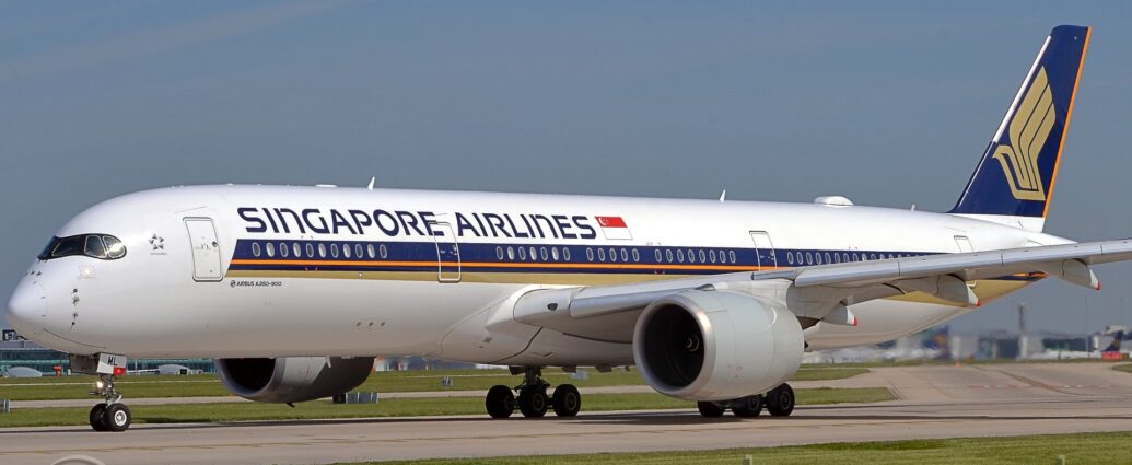 Side view of a twin engined jet airliner taxiing from right to left on a grey runway. The plane is largely white, with a purple, orange, and gold stripe running along the body from just behind the cockpit, with the gold section at the bottom getting slightly wider as it moves backwards, running under the rear fuselage. There are purple "Singapore Airlines" titles on the upper forward fuselage, next to a large Singapore" flag. The tail is purple, with a stylised golden flying bird with wings extended, and an orange stripe on the rear of the tail. Green grass fills the foreground, lining the side of the runway, with a large terminal building in the background, visible under the plane, with several planes parked next to it. Pale blue sky with grey smears of thin cloud fill the top half of the frame.