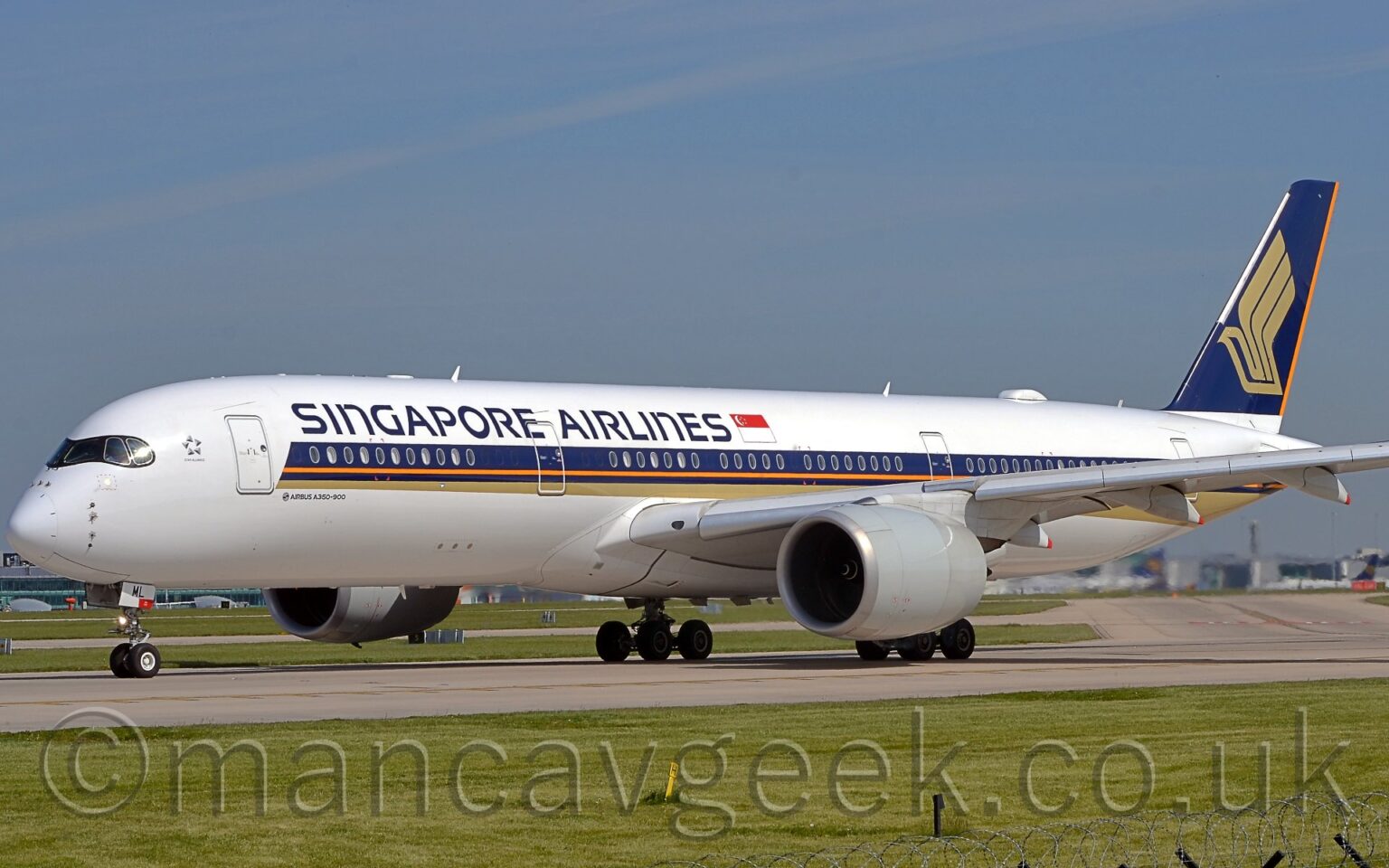 Side view of a twin engined jet airliner taxiing from right to left on a grey runway. The plane is largely white, with a purple, orange, and gold stripe running along the body from just behind the cockpit, with the gold section at the bottom getting slightly wider as it moves backwards, running under the rear fuselage. There are purple "Singapore Airlines" titles on the upper forward fuselage, next to a large Singapore" flag. The tail is purple, with a stylised golden flying bird with wings extended, and an orange stripe on the rear of the tail. Green grass fills the foreground, lining the side of the runway, with a large terminal building in the background, visible under the plane, with several planes parked next to it. Pale blue sky with grey smears of thin cloud fill the top half of the frame.