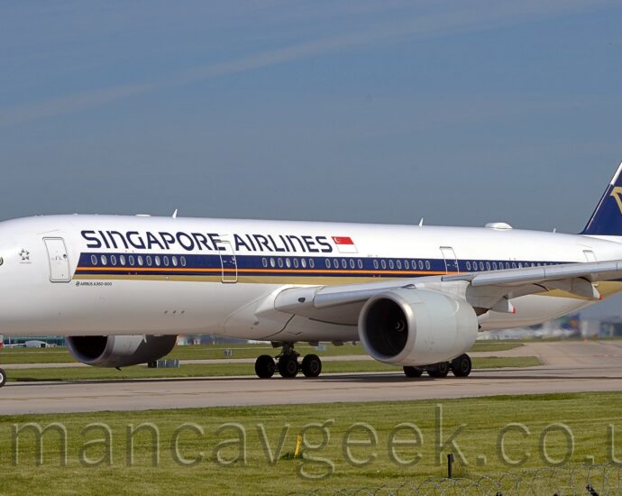 Side view of a twin engined jet airliner taxiing from right to left on a grey runway. The plane is largely white, with a purple, orange, and gold stripe running along the body from just behind the cockpit, with the gold section at the bottom getting slightly wider as it moves backwards, running under the rear fuselage. There are purple "Singapore Airlines" titles on the upper forward fuselage, next to a large Singapore" flag. The tail is purple, with a stylised golden flying bird with wings extended, and an orange stripe on the rear of the tail. Green grass fills the foreground, lining the side of the runway, with a large terminal building in the background, visible under the plane, with several planes parked next to it. Pale blue sky with grey smears of thin cloud fill the top half of the frame.