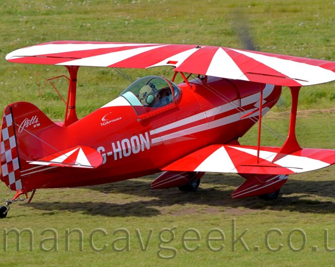 Side view of a small, single-engined biplane light aircraft taxiing from left to right on a grass airfield. The plane is mostly red, with a pair of white stripes running along the body, overlaid with the white registration "G-HOON" on the rear fuselage. White text on the tail reads "Pitts", in a rather ornate font, with smaller "S-1S" text in a more normal typeface below. The rear, moving portion of the tail is painted in a red and white checkerboard pattern. The upper surfaces of the wings and horizontal stabiliser are covered in a red and white sunburst pattern. The plane's pilot can be seen through the close, blue-tinted cockpit canopy, wearing a grey cloth flying helmet. Short green grass fills the foreground, with slightly longer grass filling the background.