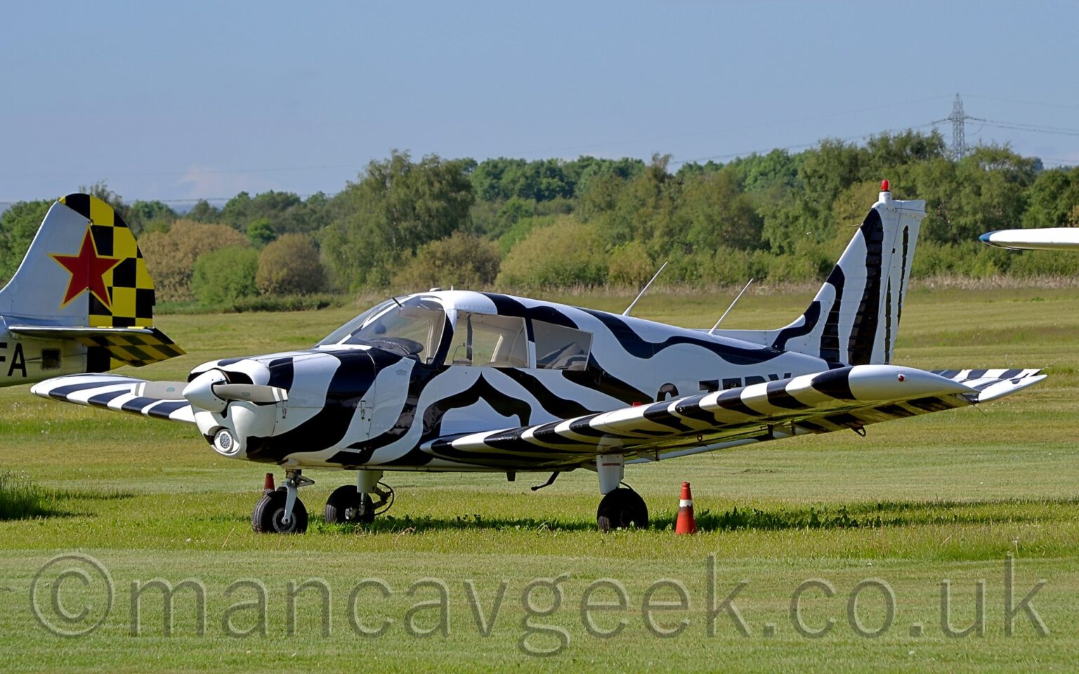 Side view of a single engined light aircraft parked facing to the left on grass. The plane is covered in wavy black and white stripes, looking vaguely like a zebra would if it had wings. Bright green grass fills the foreground and much of the background, with trees in the distance under blue skies. The tail of another light aircraft can be seen in the background on the left, silver at the front and a black and yellow checkerboard pattern at the rear, overlaid with a red star outlined in yellow, and the black letters "FA" under the horizontal stabiliser.