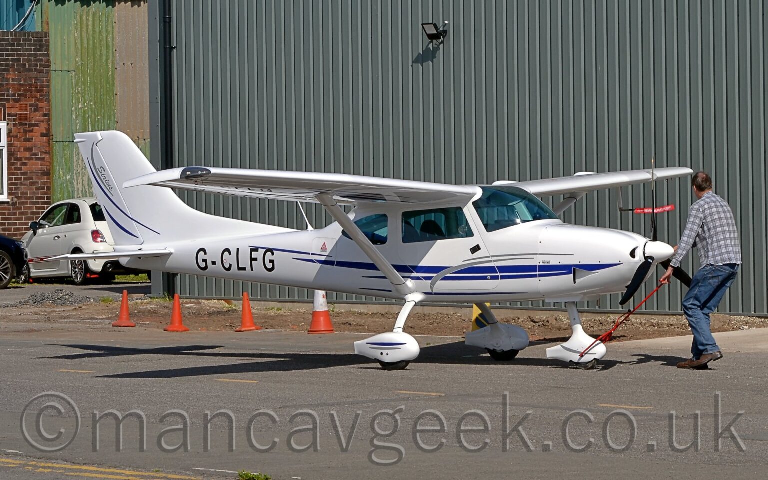 Side view of a single-engined, high-winged light aircraft being pushed around by a man wearing a check grey shirt and blue jeans. His right hand is holding the planes propellor, while his right hand is guiding a red pole that attaches to the planes nosewheel. The plane is mostly white, with a blue stripe running along the lower body, and the black registration "G-CLFG" on the sides of the rear fuselage. Black tarmac fills the foreground, with a corrugated grey metal hangar filling the background.