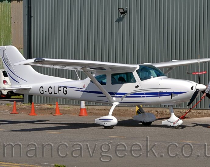 Side view of a single-engined, high-winged light aircraft being pushed around by a man wearing a check grey shirt and blue jeans. His right hand is holding the planes propellor, while his right hand is guiding a red pole that attaches to the planes nosewheel. The plane is mostly white, with a blue stripe running along the lower body, and the black registration "G-CLFG" on the sides of the rear fuselage. Black tarmac fills the foreground, with a corrugated grey metal hangar filling the background.