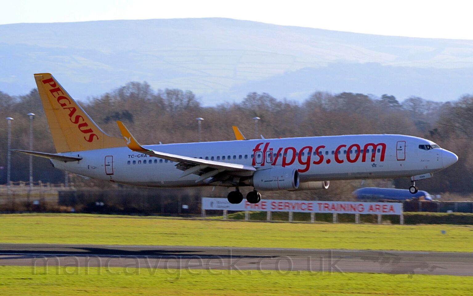 Side view of a white, twin engined jet airliner with red billboard-style "FlyPGS[dot]com" titles on the forward fuselage, yellow upturned wingtips with white winged horses in the middle, and a yellow tail with red "Pegasus" titles. The plane is flying from left to right at a very low altitude, about to land on the black runway. Green grass fills the foreground, either side of the runway, with a white sign with red text "Fire training area" in front of a black fence. The blue fuselage of a plane and trees fill the rest of the background, under grey skies.