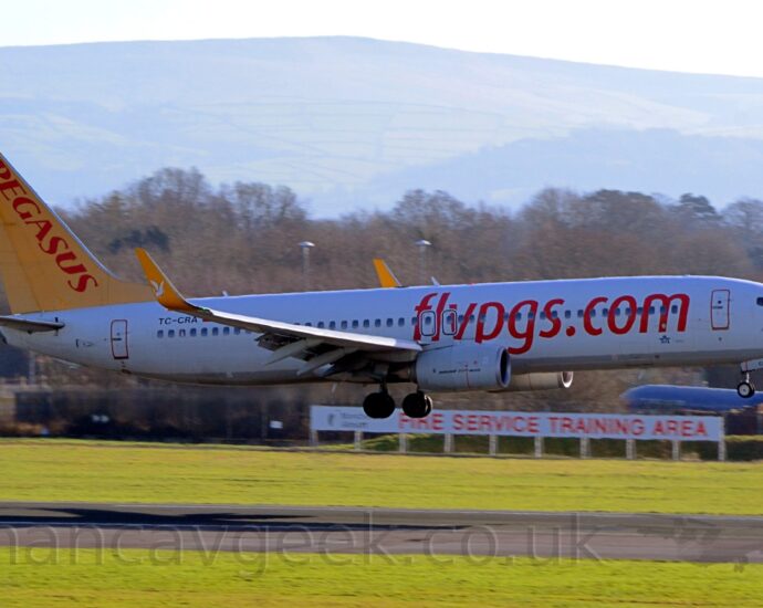 Side view of a white, twin engined jet airliner with red billboard-style "FlyPGS[dot]com" titles on the forward fuselage, yellow upturned wingtips with white winged horses in the middle, and a yellow tail with red "Pegasus" titles. The plane is flying from left to right at a very low altitude, about to land on the black runway. Green grass fills the foreground, either side of the runway, with a white sign with red text "Fire training area" in front of a black fence. The blue fuselage of a plane and trees fill the rest of the background, under grey skies.