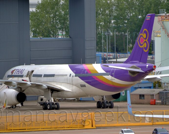 Side view of a white, twin engined jet airliner parked facing to the left and slightly away from the camera. The plane is mostly white, with purple, magenta, gold, and white diagonal bands around the rear fuselage, and a purple tail with a stylised gold and purple lotus flower in the middle. There are purple "Thai" titles next to a smaller version of that lotus logo on the upper forward fuselage. There is a set of white mobile airstairs parked by the open mid-cabin door. A yellow metal barrier stretches across the bottom of the image, with the bit on the right split down the middle so it can retract back to allow access. The wings of another couple of airliners are at the bottom of the frame, a seriously curved up-turned white wing-tip with a green, yellow, and red triangle on the outer face on the left, and a blue, somewhat straighter up-turned wingtip with a smiley face logo coming in from the right. A large grey hangar dominates the left of the image, with a duck-egg blue fuel storage tank on the right, with trees in the background behind them.