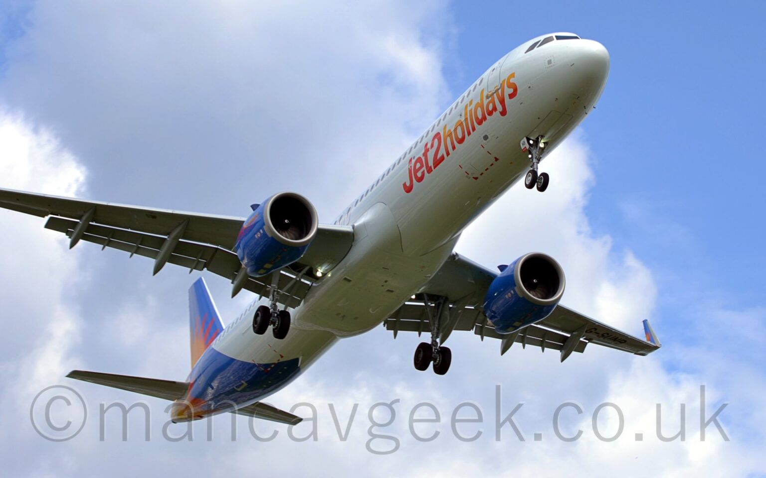 A white, twin engined jet airliner flying over head from left to right at a very low altitude. It has it's undercarriage lowered, and flaps extended from the rear of the wings, suggesting it is about to land. There are red and orange "Jet2 Holidays" titles on the lower forward fuselage, and a red and orange sunburst on the blue tail and rear fuselage, as well as on the sides of the engine pods under the wings. Blue sky with fluffs of white cloud fill the background.