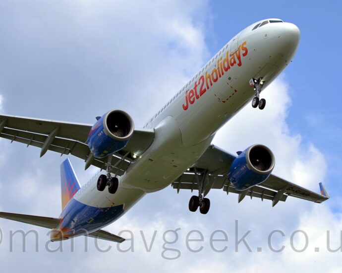 A white, twin engined jet airliner flying over head from left to right at a very low altitude. It has it's undercarriage lowered, and flaps extended from the rear of the wings, suggesting it is about to land. There are red and orange "Jet2 Holidays" titles on the lower forward fuselage, and a red and orange sunburst on the blue tail and rear fuselage, as well as on the sides of the engine pods under the wings. Blue sky with fluffs of white cloud fill the background.