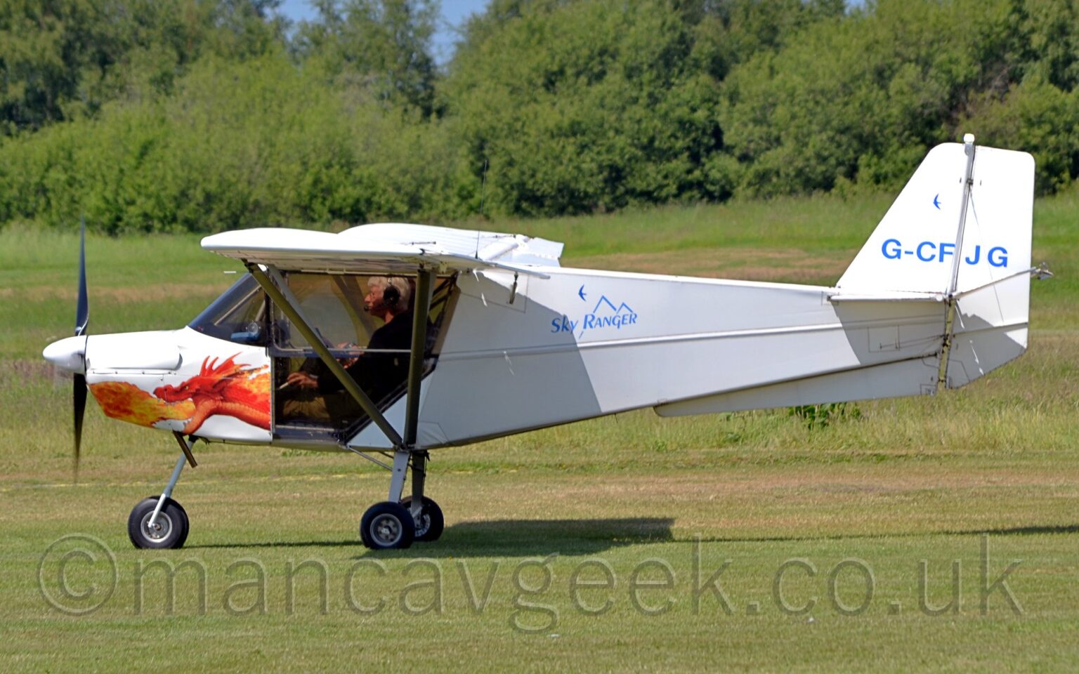 Side view of a very small, high-winged, single-engined light aircraft taxiing from right to left on grass. The plane is almost entirely white, with blue "Sky Ranger" text over a stylised image of a mountain range on the upper fuselage immediately behind the cockpit doors, the blue registration "G-CFJG" on the tail, and the head of a fire-breathing dragon that, coincidentally, lines up perfectly with the pilots legs, as seen through the bottom of the glazed doors. Green grass fills the foreground and most of the background, with trees lining the airfield perimeter in the distance, and patches of blue sky visible in gaps.