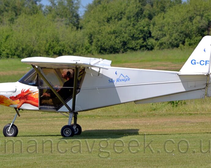 Side view of a very small, high-winged, single-engined light aircraft taxiing from right to left on grass. The plane is almost entirely white, with blue "Sky Ranger" text over a stylised image of a mountain range on the upper fuselage immediately behind the cockpit doors, the blue registration "G-CFJG" on the tail, and the head of a fire-breathing dragon that, coincidentally, lines up perfectly with the pilots legs, as seen through the bottom of the glazed doors. Green grass fills the foreground and most of the background, with trees lining the airfield perimeter in the distance, and patches of blue sky visible in gaps.