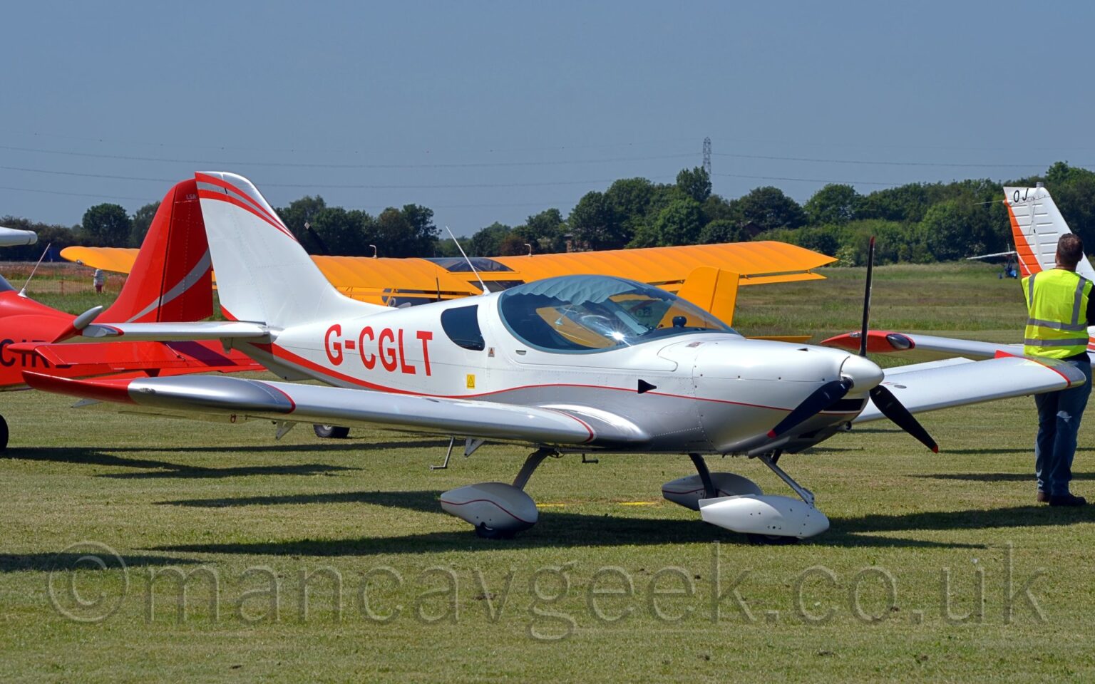 Side view of a white, single-engined light aircraft parked facing to the right on grass. The plane looks very sleek, with smooth flowing lines, and a large bubble canopy over the side-by-side cockpit. The plane has a grey belly, with a thin red stripe separating that from the white, and red registration "G-CGLT" on the side of the rear fuselage. The fixed, nose-wheel undercarriage has white sponsons over the wheels. Green grass fills the foreground, with a strong shadow being cast on the ground, suggesting the sun is high in the sky. More light aircraft can be seen in the background - a red one on the left, a yellow one with a high wing in the middle, and a white and orange one on the right. Trees in the distance meet blue sky.