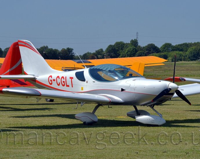 Side view of a white, single-engined light aircraft parked facing to the right on grass. The plane looks very sleek, with smooth flowing lines, and a large bubble canopy over the side-by-side cockpit. The plane has a grey belly, with a thin red stripe separating that from the white, and red registration "G-CGLT" on the side of the rear fuselage. The fixed, nose-wheel undercarriage has white sponsons over the wheels. Green grass fills the foreground, with a strong shadow being cast on the ground, suggesting the sun is high in the sky. More light aircraft can be seen in the background - a red one on the left, a yellow one with a high wing in the middle, and a white and orange one on the right. Trees in the distance meet blue sky.