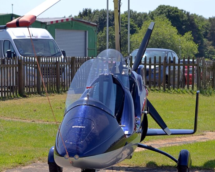 Front view of a blue, 2 seat gyrocopter parked facing the camera. The aircraft has no wings, but instead a rotor like a helicopter, but also has a pusher propellor at the rear. The fuselage is dark blue on the top surfaces, with a creamy white belly. It is parked on a small concrete slab surrounded by grass, with a brown wooden fence separating the airfield from parked cars, with tall trees behind, under pale grey skies.