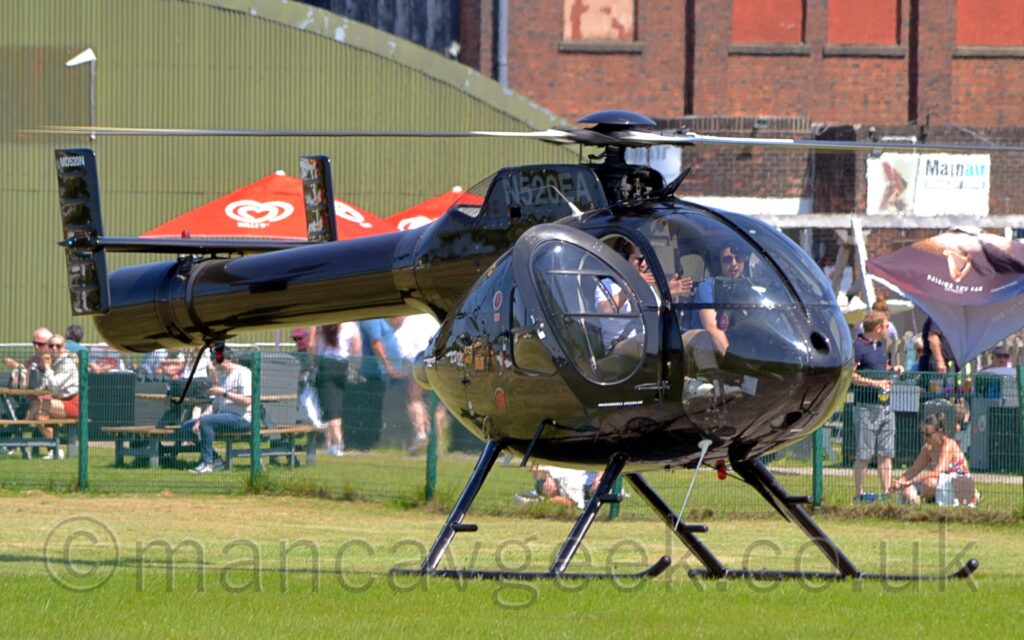 Front side view of a glossy black helicopter parked facing to the right on grass, with the motor slowly spinning and the pilots door open. The grey registration "N520EA" is on the side of the engine pods on top of the fuselage, and grey text "MD520N" is on the top of the twin tails at the end of the long, fat tail boom. Notably, there is no vertical rotor at the end of the tail boom. There are 2 people sitting in the heavily glazed cockpit, the person sitting in the right-hand seat wearing a white shirt, and using their hands to demonstrate something to the person in the left hand seat, who is wearing a blue shirt. Green grass fills the foreground, with people sitting or standing behind a green metal mesh fence in the background, and a curved, green corrugated metal hangar behind them on the left, and a large brown brick building on the right. Pretty much everyone in the photo is wearing sunglasses, so it is probably a sunny day.